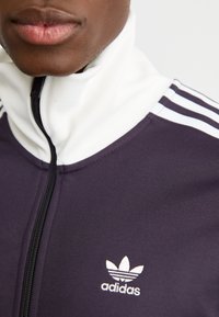 Mornarska trenirka s visokim bijelim ovratnikom, zatvaračem sprijeda i tri bijele pruge na svakom ramenu. Sadrži Adidas logo u bijeloj boji.