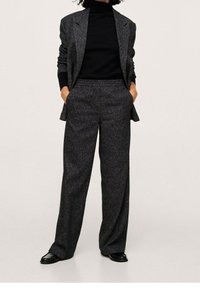 Grijs textuurpak met een op maat gemaakte blazer en wijde broek, gecombineerd met een zwarte turtleneck. Zwarte schoenen maken de look compleet.