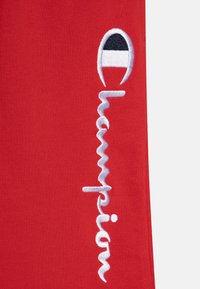 Röd tyg med broderad "Champion" logotyp och en cirkulär accent i svart, vitt och rött. Slät textur, modern design.