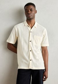 Camicia a maniche corte con bottoni di colore crema, realizzata in rete testurizzata, con taschino sul petto, bottoni marroni e colletto alla francese.