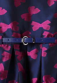 Tessuto blu navy con un motivo di fiocchi rosa, abbinato a una cintura blu uniforme con una fibbia rotonda in argento. È visibile una tessitura a rilievo.