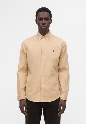 Jeune homme portant une chemise beige et blanche à rayures à manches longues boutonnée et un pantalon foncé, debout devant un fond uni.