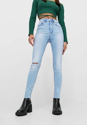Kobieta ubrana w jasnoniebieskie, wysokie spodnie skinny jeans z dziurami, zielony długi rękaw, kopertowy krótki top oraz czarne botki na obcasie, stojąca na jednolitym tle.