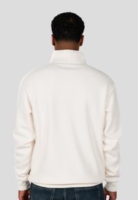 Pullover bianco a coste con colletto alto. La vista di spalle mostra maniche lunghe e una vestibilità morbida, abbinato a jeans scuri. Design semplice.