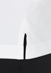 Chemise en coton blanc avec fentes latérales à l'ourlet. Texture douce, design simple et détails cousus le long des bords. Tissu noir visible en dessous.