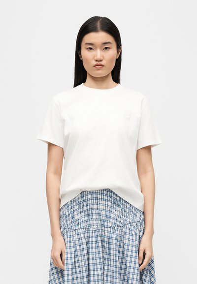 Femme aux longs cheveux bruns portant un t-shirt blanc uni à manches courtes et une jupe à carreaux bleue et blanche, faisant face à l'avant sur un fond neutre.