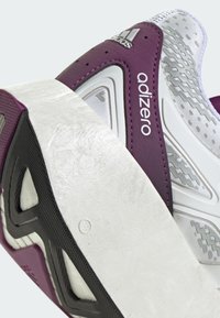 Adidas Adizero tenisice s ljubičastim sintetičkim gornjim dijelom, bijelim srednjim dijelom i crnim potplatom. Sadrže geometrijske uzorke i teksturirane akcentne elemente.