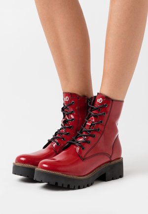 Rød læderstøvle med snørebånd, skinnende finish, sorte snørebånd, forstærket tå og tyk sort gummisål med en grov tekstur.