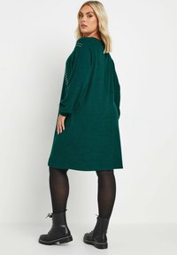 Abito maglione verde oversize, caratterizzato da un effetto tessuto a maglia, maniche lunghe e dettagli borchiati sulle spalle, abbinato a stivali militari neri e collant.