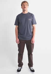 T-shirt gris à manches courtes avec une poche ornée d'un logo coloré ; associé à un pantalon marron et des baskets noires à semelles blanches.