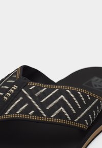 Μαύρο σανδάλι με φαρδιά υφασμάτινη λουρίδα που διαθέτει μπεζ και λευκά ζιγκ ζαγκ και πουά σχέδια και μια μικρή ετικέτα "REBEL".