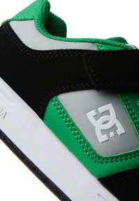 Sapato desportivo preto e verde com um upper em camurça preta texturizada, painel lateral sintético branco e um logo proeminente, com um colarinho acolchoado verde.