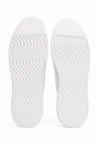 Semelles de chaussures en caoutchouc blanc avec une surface lisse et un motif de vagues texturé pour une meilleure adhérence. Détail de logo circulaire près du talon.