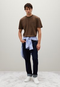 T-shirt a maniche corte marrone con tasca, camicia a righe blu legata in vita, jeans in denim scuro e sneaker bianche su uno sfondo minimale.