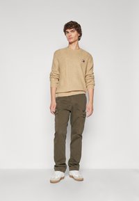 Pull en maille beige avec un petit logo sur la poitrine, associé à un pantalon cargo vert olive avec des poches zippées. Baskets blanches.