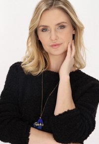 Schwarzer, strukturierter Pullover; goldene Kettenhalskette mit einem blauen Quasten und Perlen. Das Model posiert mit einer Hand im Gesicht, natürlicher Hintergrund.