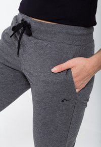 Dorosła osoba nosząca ciemnoszare dresowe spodnie z sznurkiem, z ręką w kieszeni, oraz czarny top, pokazana od talii do uda.