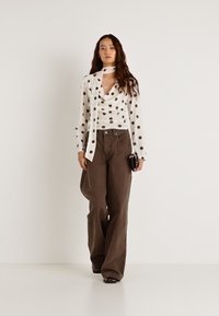 Biała bluzka z długim rękawem w brązowe groszki, z dekoltem w draperię, do tego luźne brązowe spodnie, w ręku czarna kopertówka.