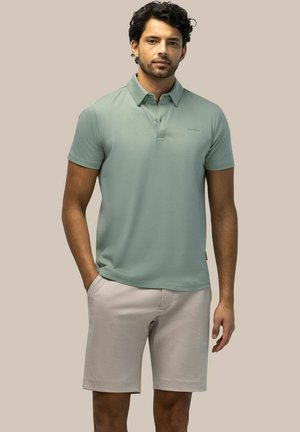 Man with curly hair wearing a light green short-sleeve polo shirt and beige shorts standing with one hand in pocket against plain background.