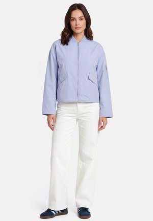 Frau trägt hellblaue Jacke mit Reißverschluss, weiße Hosen mit weitem Bein und marineblaue Turnschuhe, steht vor einem schlichten weißen Hintergrund.