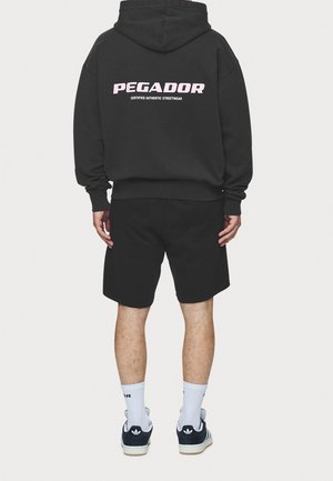Mężczyzna w czarnej bluzie z kapturem z napisem "PEGADOR Certified Authentic Streetwear", czarnych szortach, białych skarpetkach i czarnych butach sportowych Adidas, widziany od tyłu.