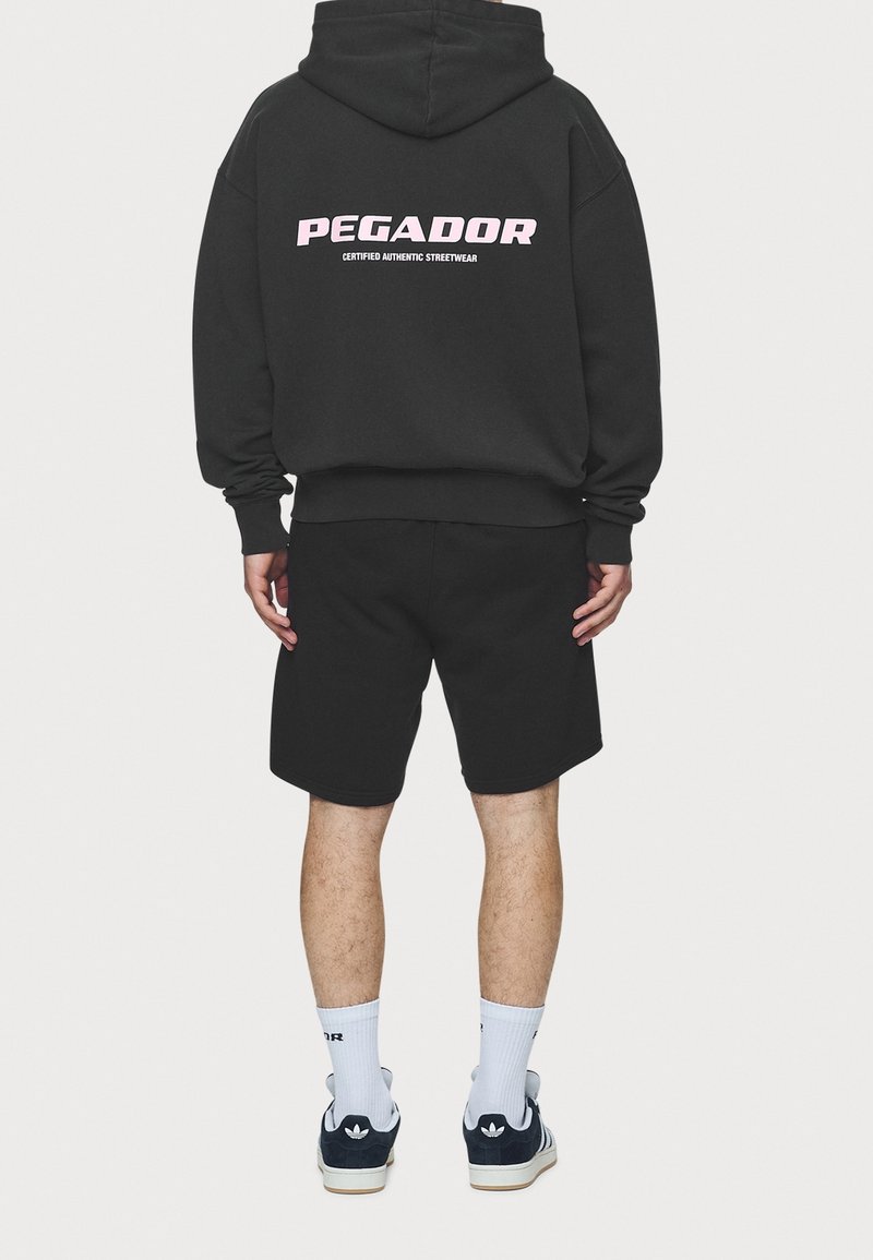 Homme portant un sweat à capuche noir avec le texte "PEGADOR Certified Authentic Streetwear", short noir, chaussettes blanches et baskets Adidas noires, vu de dos.