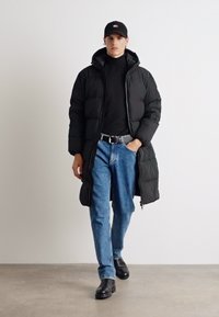 Black padded long coat with a hood, black turtleneck, blue jeans, and black ankle boots, featuring a sleek, minimalist design.