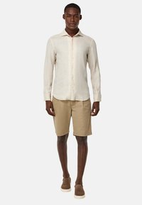 Camicia in lino beige chiaro con bottoni e colletto, abbinata a pantaloni corti in cotone color sabbia e scarpe marroni slip-on. Un outfit semplice e casual.