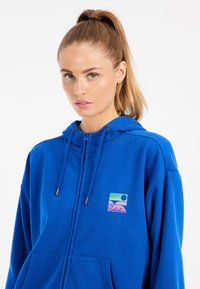 Sudadera con cierre en azul royal, capucha con cordón, bolsillo canguro y logotipo frontal que representa un paisaje con colores pastel y el texto "Protest".