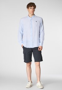 Giovane uomo che indossa una camicia azzurro chiaro a maniche corte, pantaloni cargo scuri e sneakers bianche, in piedi su uno sfondo neutro.
