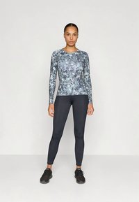 Langärmelig sporttop in een grijs-blauw camouflagepatroon, gecombineerd met zwarte leggings en zwarte sportschoenen.