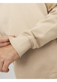 Felpa beige con polsini a coste e morbida texture del tessuto; mani che regolano le maniche, mostrando dettagli di cucitura.