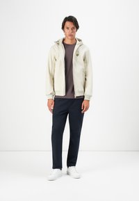 Felpa leggera con zip di colore chiaro, con un logo sul petto, abbinata a una maglietta scura e pantaloni blu scuro. Sneakers bianche completano l'outfit.