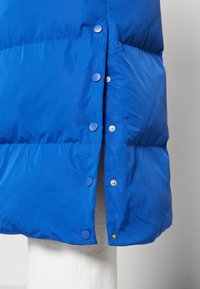 Blå pufferjakke med en glatt, skinnende tekstur, trykknaplukking og avrundet nederkant. Stoffet virker lett og varmt.