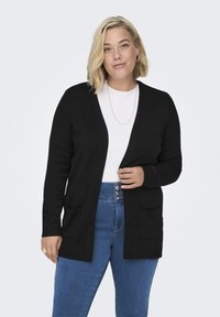Zwarte cardigan met een v-halsontwerp, geribde manchetten en twee voorzakken. Draagt over een witte, textuurshirt en blauwe jeans.