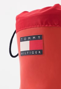 Punainen kuminen sadetakki mustalla ja valkoisella Tommy Hilfiger -logopatchilla ja mustalla vetoketjulla valkoista taustaa vasten.