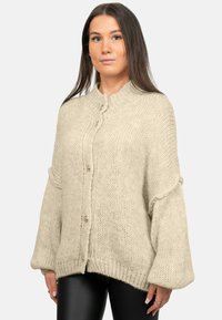 Cardigan lavorato a maglia beige con texture a coste, chiusura a grande bottone e maniche ampie. Il tessuto è morbido e leggermente oversize nel fitting.