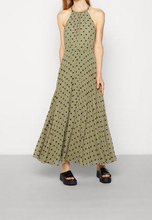 Mujer vistiendo un vestido largo de cuello halter color verde oliva con lunares negros y sandalias de plataforma negras, de pie contra un fondo blanco.