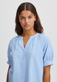 Camicia azzurra chiara con bottoni, caratterizzata da strisce bianche verticali, maniche corte a sbuffo e un design del colletto casual.