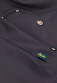 Marineblauwe waterdichte jas met een metalen drukknoopsluiting en een klein Zweeds vlaggetje op de voorkant van de zak. Gladde, duurzame textuur.