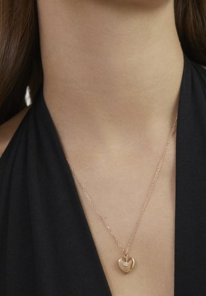 Roségold-Halskette mit einem herzförmigen Anhänger und einem kleinen dekorativen Akzent, präsentiert vor einem dunklen Stoffhintergrund.