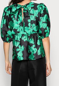Donna che indossa una blusa nera di satin con un motivo floreale verde, maniche a sbuffo e un fiocco legato sul collo della schiena, abbinata a pantaloni neri.