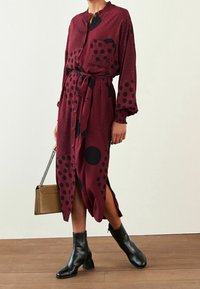 Abito bordeaux con pois neri e motivi astratti, caratterizzato da una vita legata, bottoni sul davanti e maniche lunghe. Indossato con stivali neri alla caviglia.