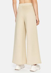 Lange beige broek met een elastische tailleband, een gladde textuur en een lichtgewicht stof, die boven de enkel eindigt. Er worden witte sneakers gedragen.