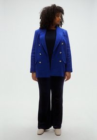 Plavi dvoredni blejzer sa zlatnim gumbima, uparen s crnim širokim hlačama i kremastim cipelama. Glatka tekstura tkanine.