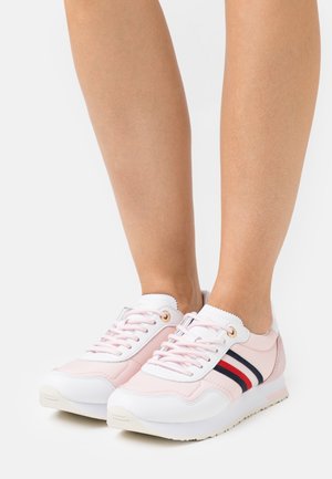 Jambes portant des baskets roses et blanches avec des rayures rouges et bleu marine, debout sur un fond blanc.