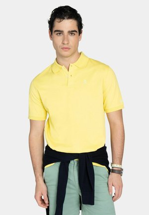 Joven vestido con una camiseta tipo polo amarilla, pantalones cortos verdes y un suéter negro atado alrededor de la cintura, posando frente a un fondo liso.