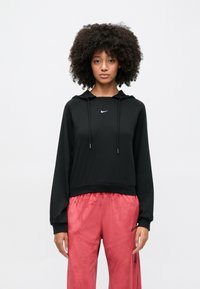 Fiatal, göndör hajú személy fekete Nike kapucnis pulóvert és élénk rózsaszín Nike edzőnadrágot visel, és egyszínű fehér háttér előtt áll.