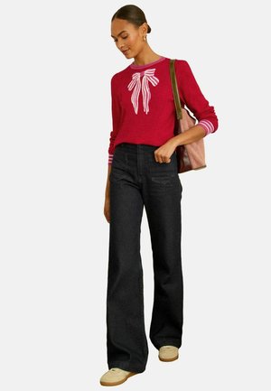 Roter Pullover mit weißem Schleifendesign, gestreiften Bündchen und hochtaillierter schwarzer ausgestellter Jeans. Braune Tasche und helle Turnschuhe vervollständigen das Outfit.
