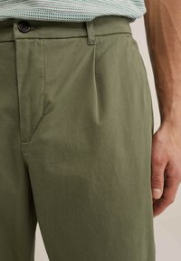 Pantalones verde oliva de tela ligera con ligera textura, presentan una cintura suave y un cierre de un solo botón, con un bolsillo de mano visible.
